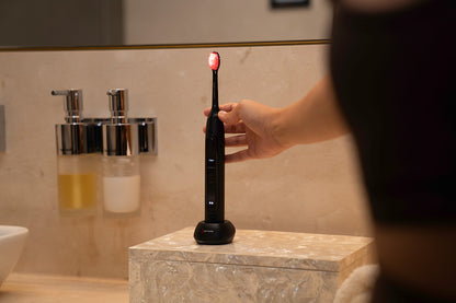 Person holding a black electric toothbrush with a red light on a bathroom counter.